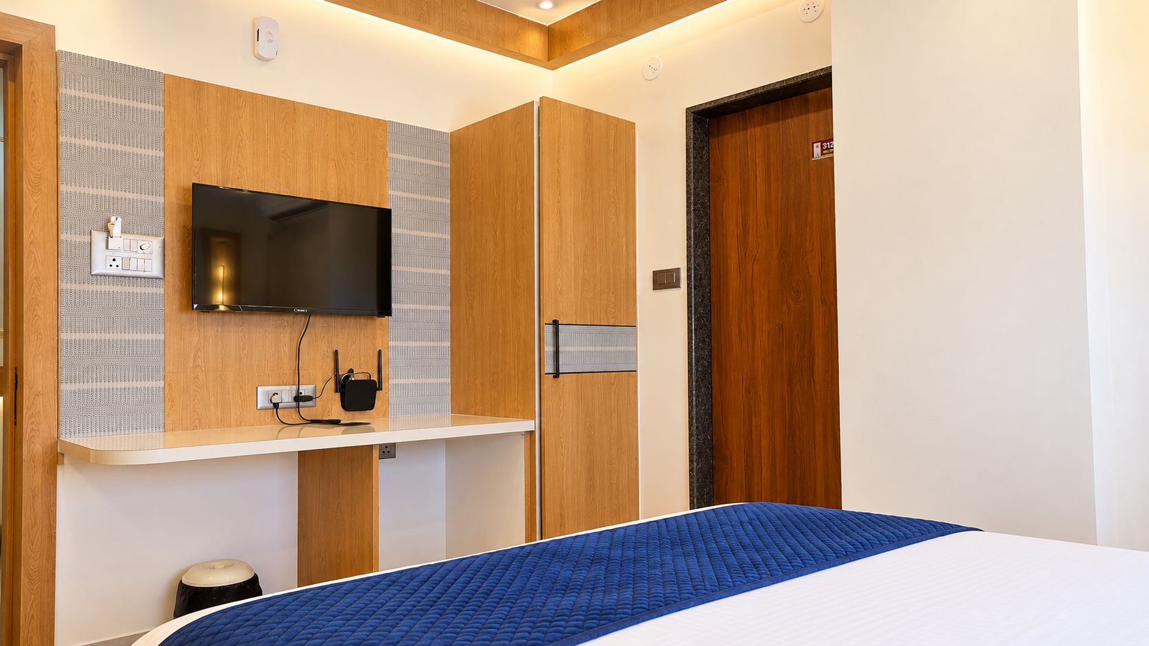 Une chambre d’hôtel comprenant un lit, un téléviseur à écran plat fixé sur un panneau en bois, une petite table de chevet avec une prise électrique et une porte dotée d’une serrure.