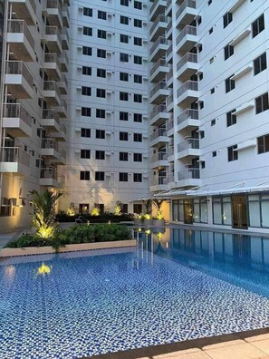 A high-rise residential building with multiple balconies, a swimming pool with a tiled floor, and landscaped areas with plants.