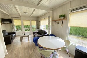 A room with a sofa, a round table, and chairs, a television mounted on the wall, and a window with a view of greenery. - Type C "Zee, Bos en Duin" (Zoutelande)