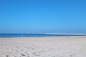 A wide sandy beach with a clear blue sky and a distant view of wind turbines and a breakwater. - Lieblingsplatz de Banjaard (Kamperland)