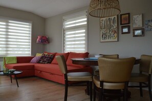 A living room with a red sofa, a round table, and chairs. There are two windows with blinds, a lamp, and framed artwork on the wall. - Cabin in Schagen with swimming pool (Schagen)