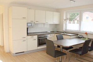 A modern kitchen with white cabinets, a dining table with chairs, and a window with curtains. - Holiday apartment with 2 bedrooms (Gronau)