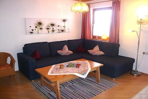 Een woonkamer met een donkergrijze bank, een houten salontafel, een rieten fauteuil, een schilderij aan de muur en een raam met gordijnen.