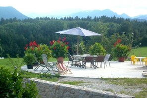 Een terras buiten, met een tafel en stoelen, omgeven door plantenpotten en een uitzicht op een bebost gebied.