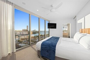 A hotel room with a large bed, a ceiling fan, a TV mounted on the wall, and a view of a cityscape through floor-to-ceiling windows. - Luxury Broadwater Retreat at Lagoon Main Beach (Main Beach)