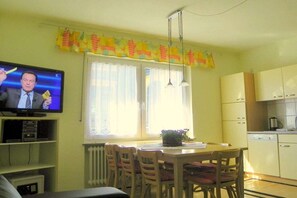 A dining area with a table and chairs, a television displaying a news program, and a kitchen area with cabinets and appliances. - Apartment with balcony overlooking the castle (Bernkastel-Kues)