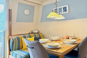Een eetgedeelte met een houten tafel gedekt voor vier personen, een bank met kussens, een klok aan de muur en een lamp boven de tafel.