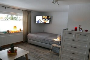 A small, well-lit room with a bed, a television mounted on the wall, a table with fruit, and a dresser with drawers. - Studio Auszeit Sauerland (59939 Olsberg, Deutschland)