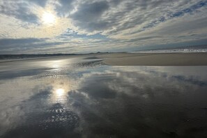 Une plage au sable humide et brillant, reflétant la lumière, et un ciel partiellement nuageux.