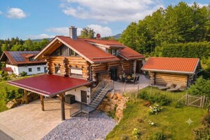 A log cabin with a red roof, a covered patio, and a stone staircase leading to a deck. - Cabin in Spiegelau with swimming pool (Spiegelau)