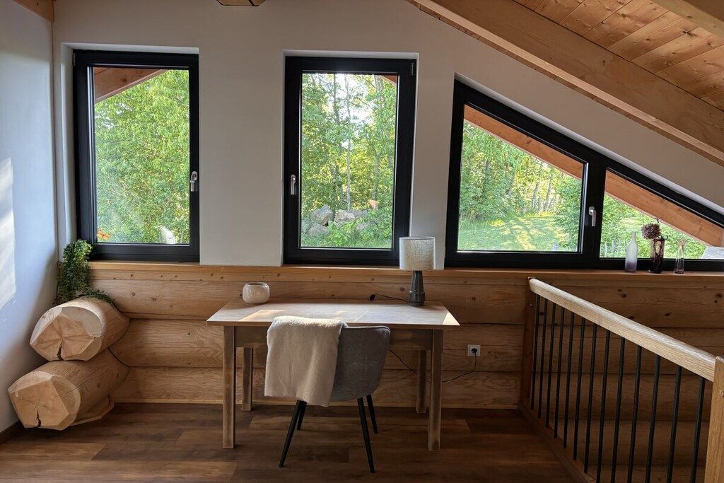 Ein Zimmer mit einem Holztisch, zwei Fenstern mit schwarzen Rahmen und Blick auf Grünfläche.