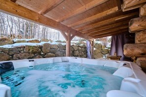 A hot tub under a wooden roof with a stone wall and trees in the background.
