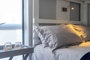 Un lit avec une literie blanche, une table de chevet sur laquelle reposent deux bocaux en verre, et une lampe fixée au mur.
