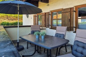 An outdoor patio with a table and chairs, a black umbrella, and wooden shutters on the windows. - Leni holiday apartment (Bischofswiesen)