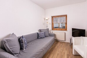 Een woonkamer met een grijze bank, een witte fauteuil, een televisie en een raam met gordijnen.