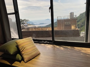 A room with a large window offering a view of a coastal landscape, a wooden bench, and a striped pillow. - M Bar House (New Taipei City)
