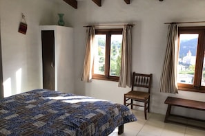 A bedroom with a bed, a chair, a bench, and two windows with curtains. - Holiday home with 2 bedrooms (Puigpunyent \/ Mallorca)