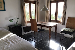 Quarto com cama, escrivaninha, cadeira, luminária e vista pela janela.