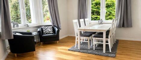 A dining area with a white table and chairs, grey curtains, and a view of greenery outside.
