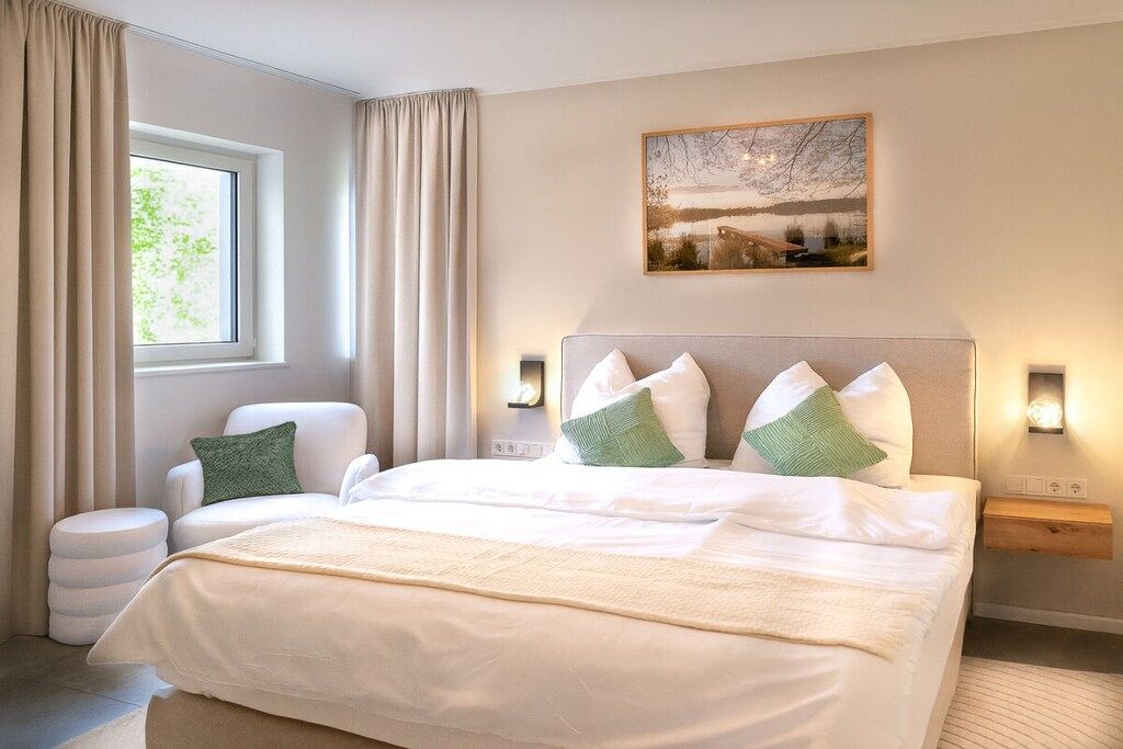 Une chambre d’hôtel avec un grand lit, une chaise, une table de chevet, une commode, une lampe fixée au mur et une fenêtre avec des rideaux.
