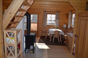 A wooden cabin interior with a dining table, chairs, a black stove, and a bookshelf. - Holiday home with 1 bedroom (Adelschlag Ortsteil  Möckenlohe)