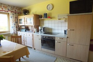 A kitchen with a stove, oven, and microwave. There is a clock on the wall and a window with curtains. - 1 - Gertraud Weiderer (Lindberg)