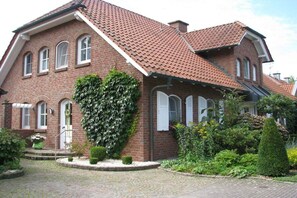 Un bâtiment en briques doté d’un toit en pente, de fenêtres en arc de cercle et d’un jardin bien entretenu, agrémenté de haies et d’arbustes.