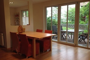 A dining area with a wooden table and red chairs, a kitchen area with a refrigerator, and a sliding glass door leading to an outdoor patio with tables and chairs. - Seedorf Hörster Heide (Warendorf)