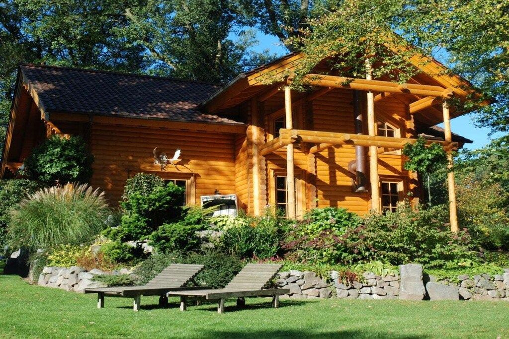 A wooden cabin with a porch, surrounded by a landscaped garden with a stone wall and wooden benches.