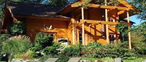 Puinen mökki, jossa on kuisti, ja sen ympärillä on maisemoitu puutarha, jossa on kivimuuri ja puisia penkkejä.
