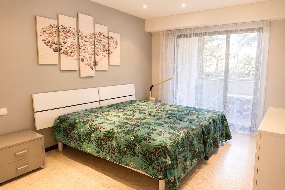 A bedroom with a bed, a nightstand, a bedside table, a lamp, and a window with curtains.
