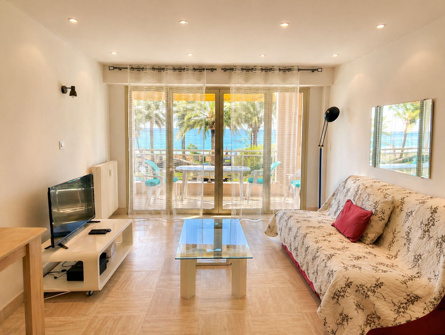 A modern living room with a glass coffee table, a sofa with a floral pattern, a TV on a stand, and a view of a balcony with outdoor furniture.