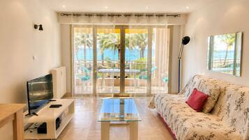 A modern living room with a glass coffee table, a sofa with a floral pattern, a TV on a stand, and a view of a balcony with outdoor furniture.
