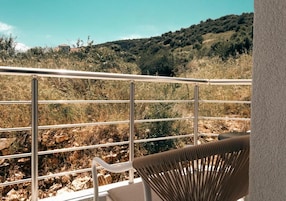 Un balcón con barandilla metálica, una silla y vistas a un paisaje natural con colinas y vegetación escasa.