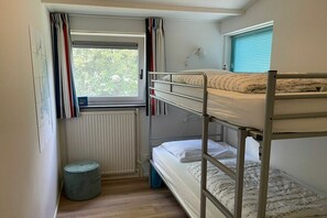 A dormitory room with bunk beds, a radiator, a window with curtains, and a small round stool. - Holiday apartment with 2 bedrooms (Ballum)