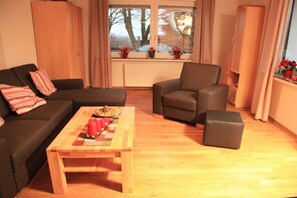 Un salon aménagé avec un canapé, un fauteuil, un pouf et une table basse sur laquelle se trouvent une couverture et une assiette.