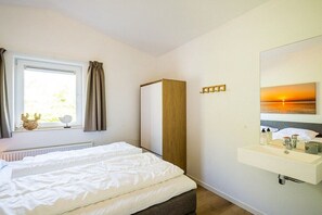 Een slaapkamer met een bed, een raam met gordijnen, een nachtkastje, een spiegel en een wandplank met toiletartikelen.