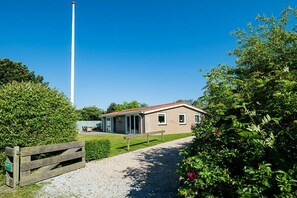 Een laag gebouw met een grindpad erheen, omgeven door groen en een houten hek.