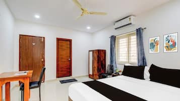 A hotel room with a bed, a desk, a chair, a ceiling fan, an air conditioning unit, and two framed artworks on the wall.