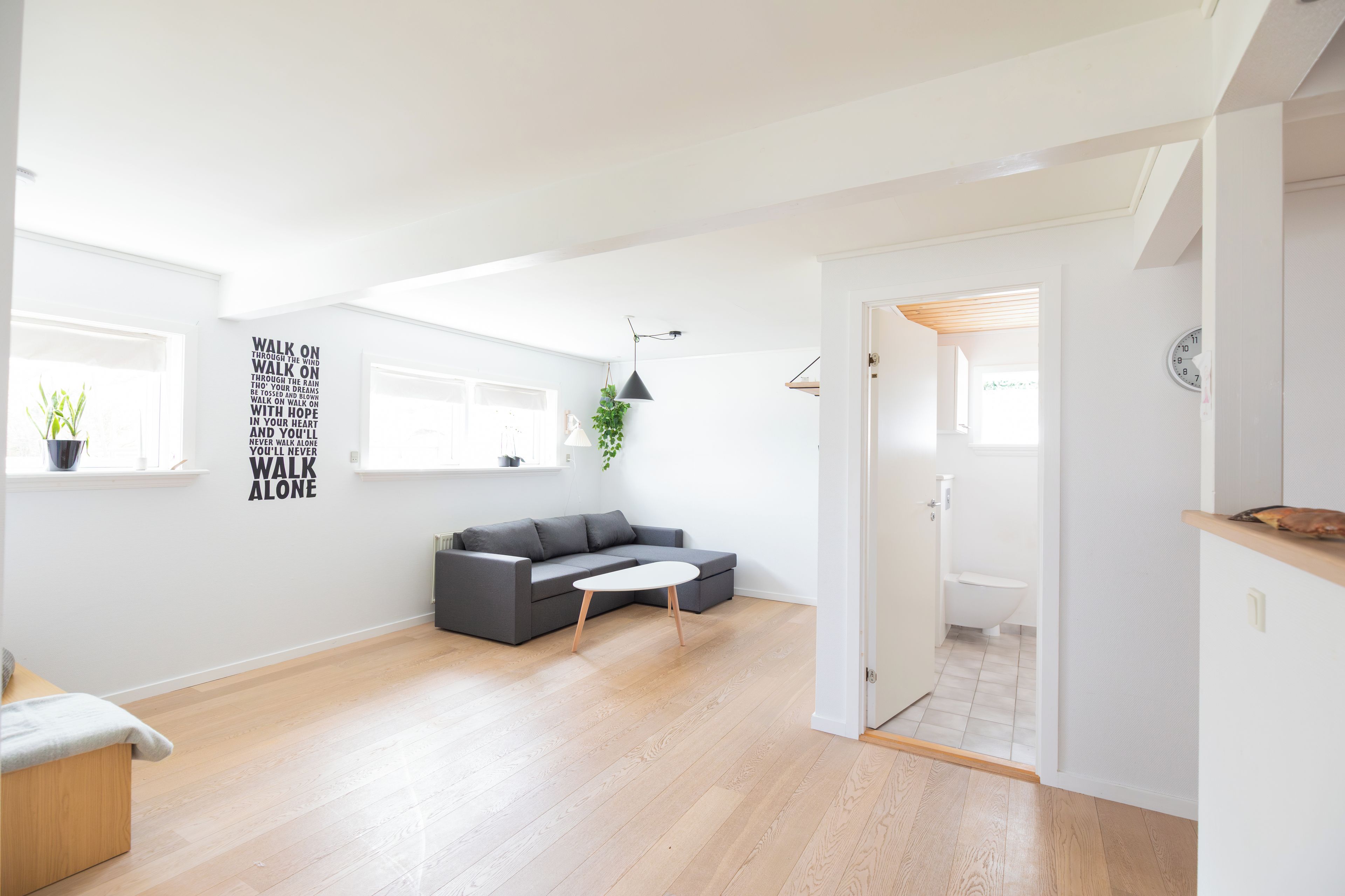 Un salon moderne avec un canapé gris, une petite table blanche, un plancher en bois et une grande fenêtre donnant sur un beau paysage.