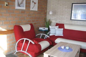 A room with a brick wall, a white sofa with red cushions, a small table with decorative items, and a framed picture above the sofa. - Beach holiday and sea (St. Maartensvlotbrug)
