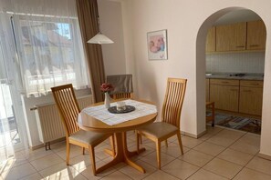 Un comedor con una mesa redonda, dos sillas, un florero con flores y una ventana con cortinas.