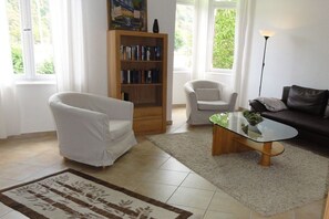 A living room with a white armchair, a sofa, a glass coffee table, a bookshelf, and a lamp. - Holiday apartment with 1 bedroom (Koblenz)