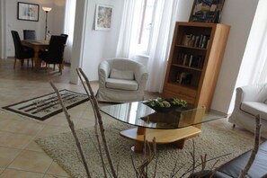 A living room with a glass coffee table, a white armchair, a bookshelf, and a dining area with chairs and a table. - Holiday apartment with 1 bedroom (Koblenz)