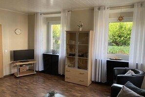 Ein Wohnzimmer mit Fernseher auf einem Schrank, einer Vitrine, einem schwarzen Sessel und einem fenster mit Vorhängen.