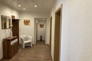 Un pasillo con suelo de madera, una silla blanca con cojín estampado y un aparador de madera con cajones.