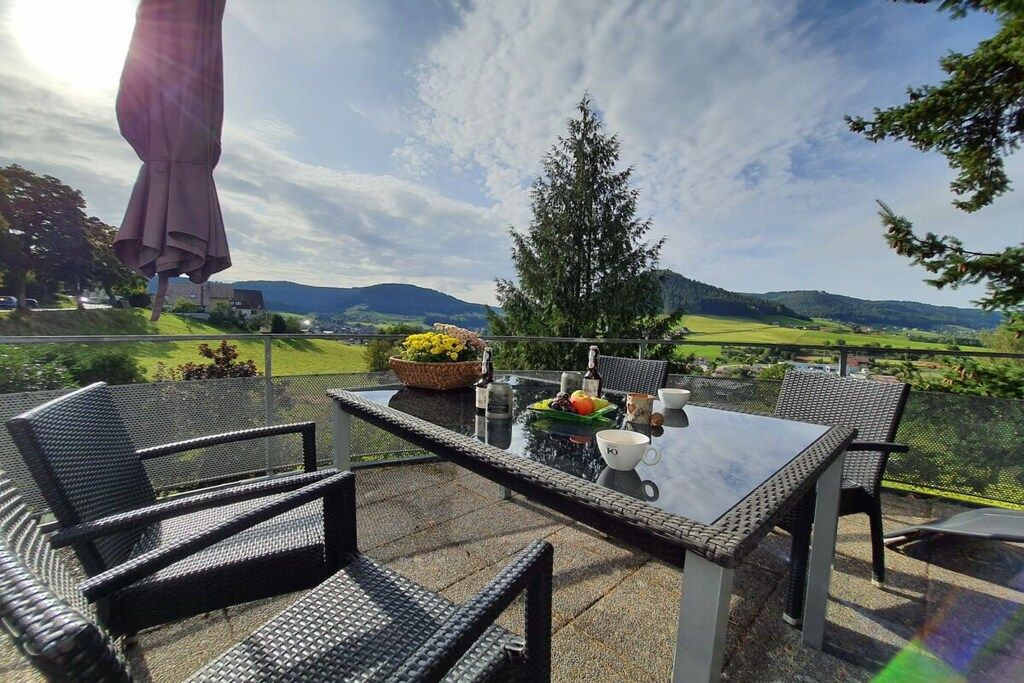 An outdoor patio with a glass table and wicker chairs, overlooking a scenic landscape with hills and trees.