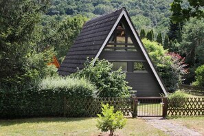 A small, dark-colored wooden cabin with a triangular roof, surrounded by dense greenery and a wooden fence. - Holiday home in Landau with swimming pool (Landau)