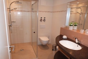 Casa de banho com chuveiro envolto em vidro, sanita branca, lavatório com espelho e um contentor de lixo.