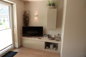 Ein modernes Wohnzimmer mit einem Flachbildfernseher, einem kleinen Tisch mit Dekoration, einem an der Wand befestigten Regal mit einer Pflanze und einem Fenster mit Blick auf ein Gebäude.
